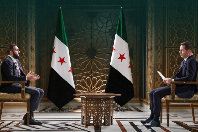 مقابلة تلفزيونية للرئيس السوري أحمد الشرع مع أحد الإعلاميين، يظهر في الخلفية علم الثورة السورية بثلاث نجوم حمراء